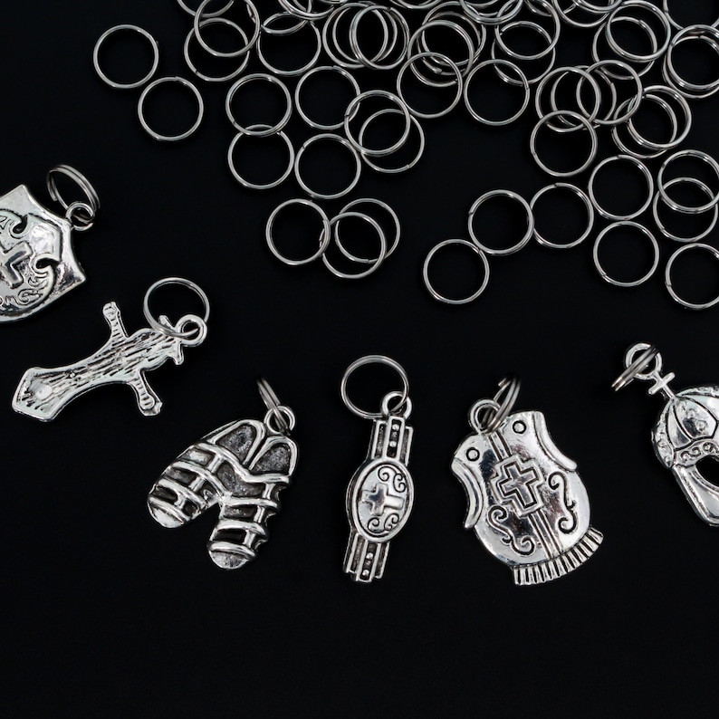 May include: A collection of silver-toned charms featuring various religious symbols, including a cross, a shield, a helmet, and a vest. The charms are arranged on a black background with a pile of silver jump rings.