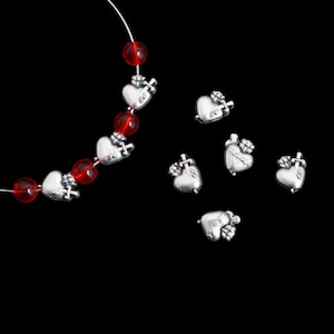 May include: A necklace with red beads and silver heart charms, each pierced by a cross. Several loose heart charms are scattered on a black background. The charms have a detailed, textured appearance.