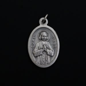 May include: Silver oval pendant with a relief image of a man in a priest's robe, with the text "CURE D'ARS" around the edge.