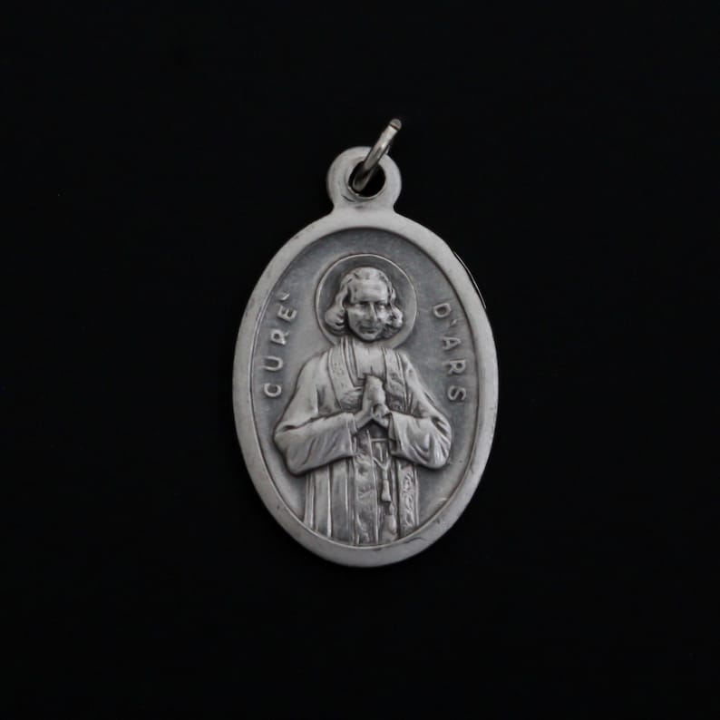 May include: Silver oval pendant with a relief image of a man in a priest's robe, with the text "CURE D'ARS" around the edge.