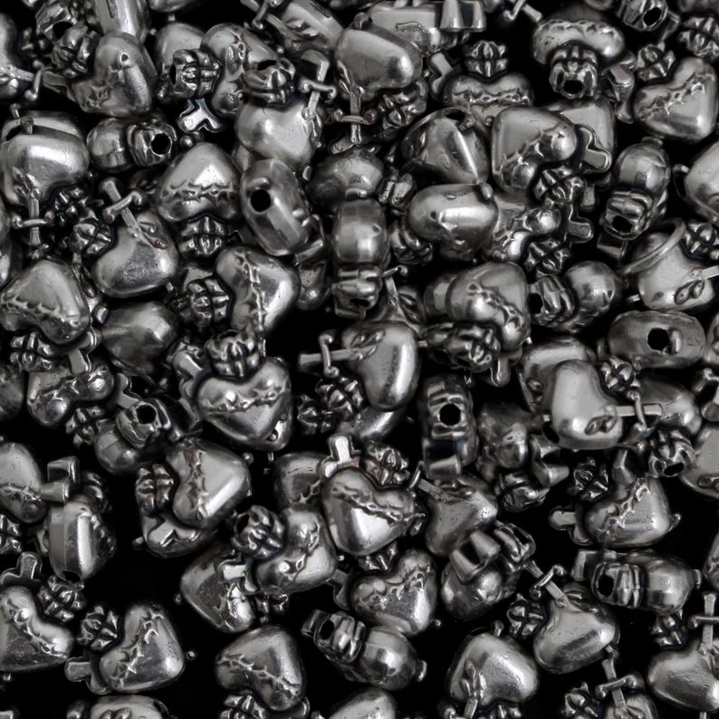 May include: A close-up shot of a pile of silver-colored heart-shaped beads. Each bead has a detailed design, including a cross and a crown. The beads are scattered, creating a textured surface.