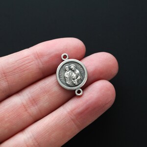 May include: A small, round, silver-toned pendant with two small holes for attachment. The pendant features a detailed relief of a family and the words "HOLY FAMILY ITALY" around the edge. The pendant is held in a person's hand against a dark background.