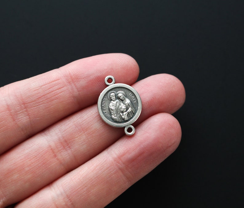 May include: A small, round, silver-toned pendant with two small holes for attachment. The pendant features a detailed relief of a family and the words "HOLY FAMILY ITALY" around the edge. The pendant is held in a person's hand against a dark background.