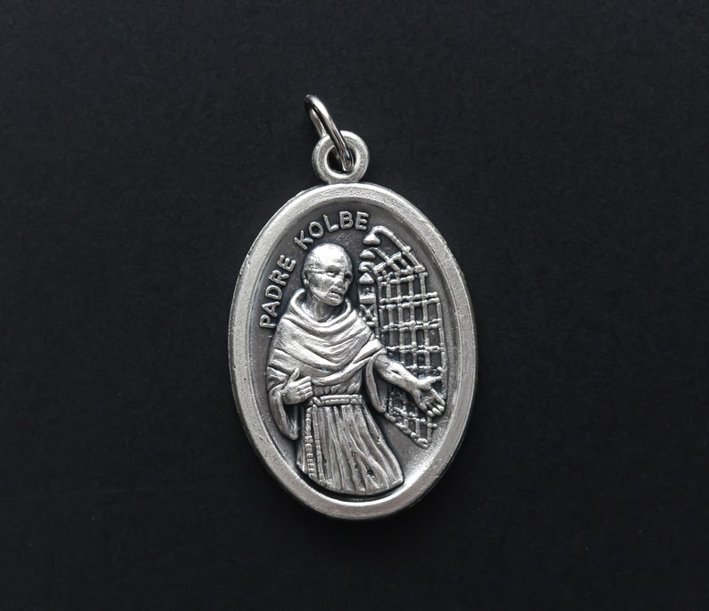 May include: Silver oval pendant with a depiction of Padre Kolbe, a Catholic saint, wearing a brown robe and holding a fence. The pendant has the words "Padre Kolbe" inscribed on it.