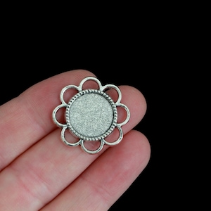 May include: A silver-toned metal flower-shaped jewelry finding with a circular recessed area for setting a cabochon. The finding has a scalloped edge and a textured surface, perfect for crafting pendants, earrings, or other jewelry pieces.