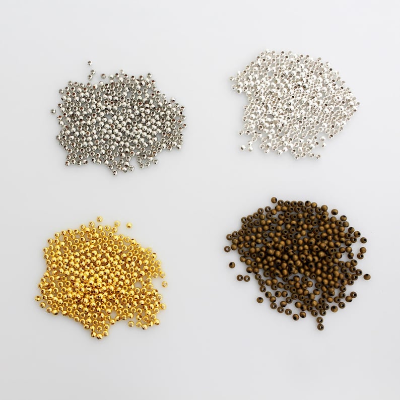 May include: Four distinct piles of small, round beads arranged in a square formation. The top left pile is metallic silver, the top right pile is bright silver, the bottom left pile is polished gold, and the bottom right pile is dark bronze.