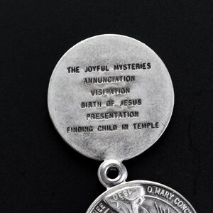May include: A silver religious pendant with the text "THE JOYFUL MYSTERIES" and other religious text. The pendant is circular with a loop for a chain. The background is black.