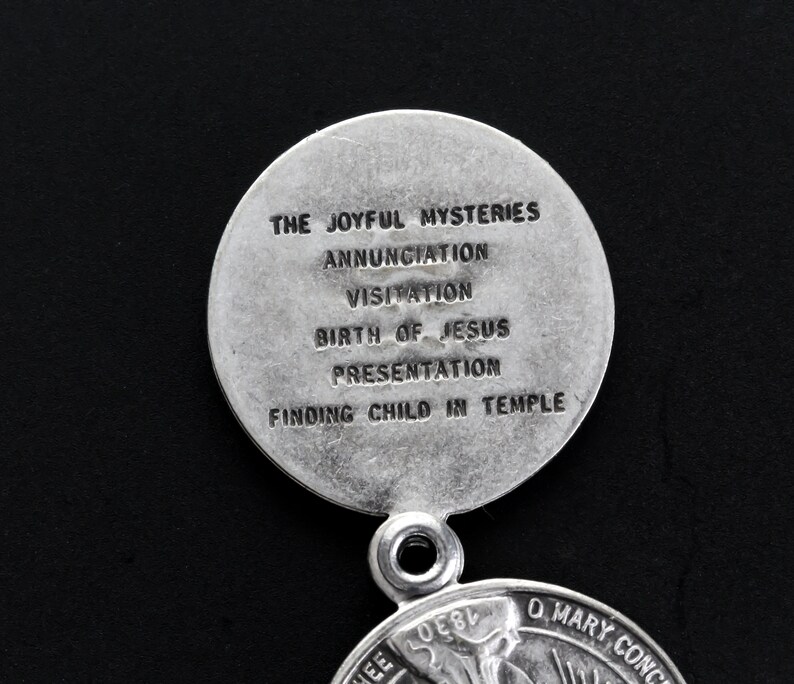 May include: A silver religious pendant with the text "THE JOYFUL MYSTERIES" and other religious text. The pendant is circular with a loop for a chain. The background is black.