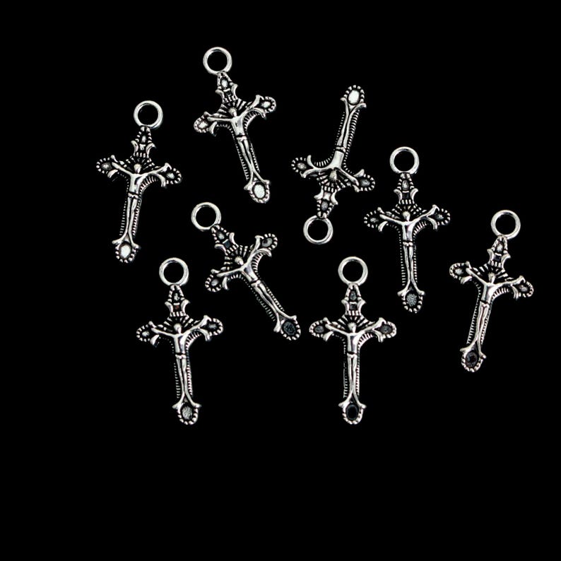 May include: A collection of eight silver-toned metal crucifix charms, each featuring a detailed figure of a person on a cross with an attached circular jump ring at the top. The charms have an oxidized finish that highlights the intricate design.