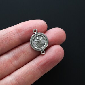 May include: A small, silver-colored pendant with a circular design. The pendant features a detailed image of a dove with radiating lines, and two small loops for attachment. The pendant is held in a hand against a black background.