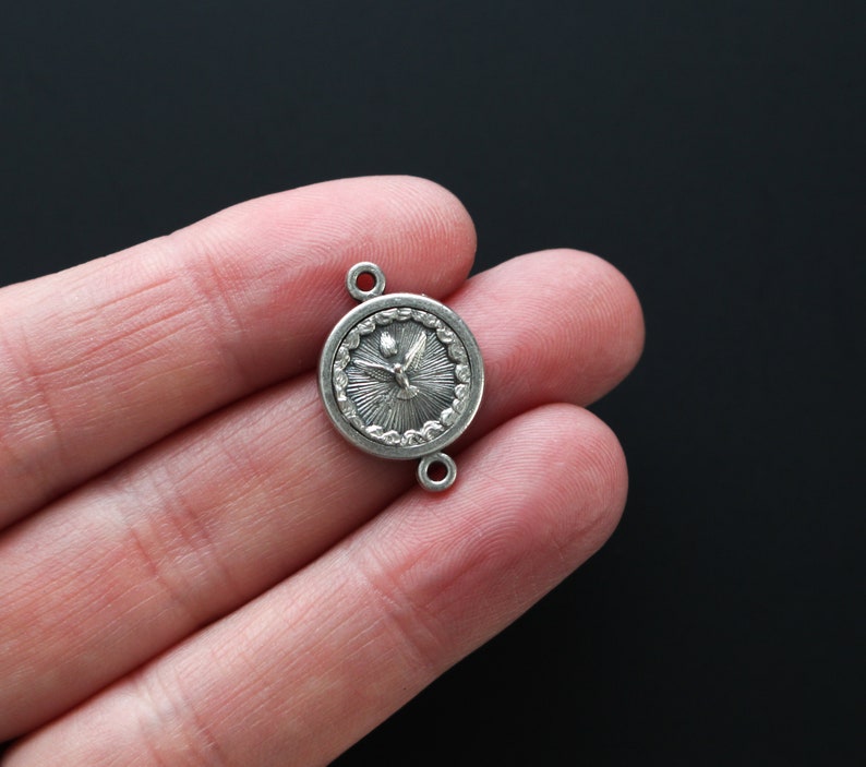 May include: A small, silver-colored pendant with a circular design. The pendant features a detailed image of a dove with radiating lines, and two small loops for attachment. The pendant is held in a hand against a black background.