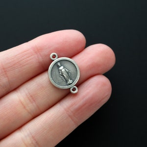 May include: Silver charm featuring a round pendant with a raised image of the Virgin Mary. The charm has two jump rings for attaching to a necklace or bracelet.