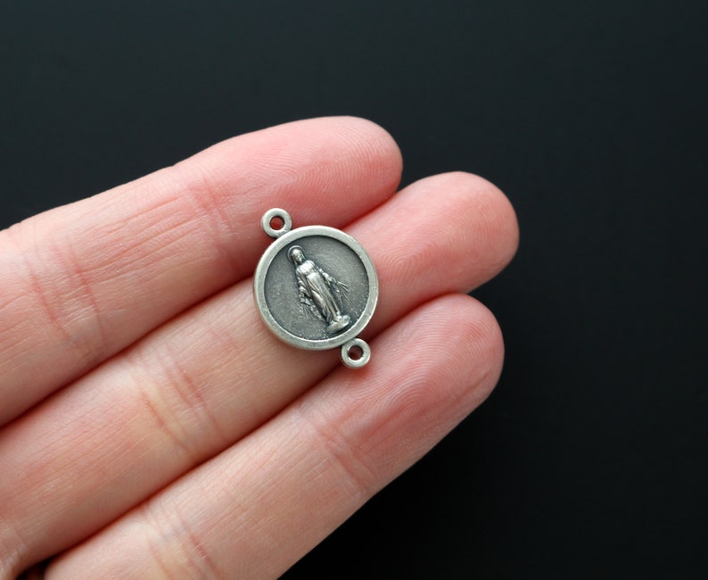 May include: Silver charm featuring a round pendant with a raised image of the Virgin Mary. The charm has two jump rings for attaching to a necklace or bracelet.