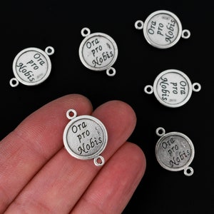 Op de afbeelding: Zilverkleurige metalen bedels met de inscriptie "Ora pro Nobis" in een cirkelvormig ontwerp. Elke bedel heeft twee kleine lussen om aan sieraden of andere knutselwerken te bevestigen. Zwarte achtergrond.