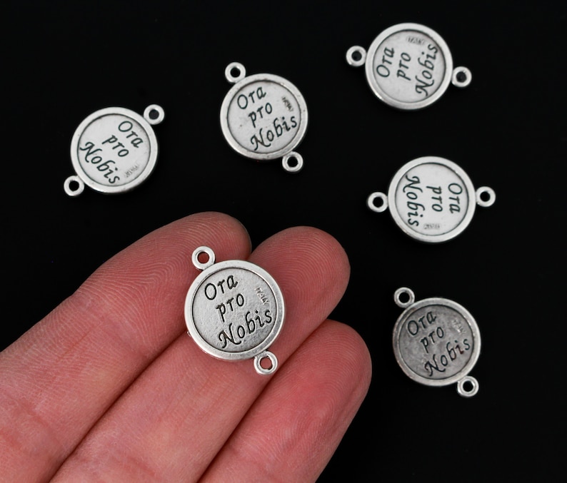 Op de afbeelding: Zilverkleurige metalen bedels met de inscriptie "Ora pro Nobis" in een cirkelvormig ontwerp. Elke bedel heeft twee kleine lussen om aan sieraden of andere knutselwerken te bevestigen. Zwarte achtergrond.