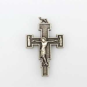 Op de afbeelding: Een zilverkleurige kruis hanger met een gedetailleerde afbeelding van Jezus Christus aan het kruis. Het kruis heeft ingewikkelde houtsnijwerk en een klein opschrift bovenaan.