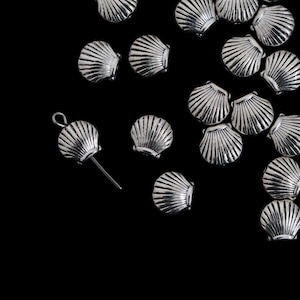 May include: Silver-colored seashell charms scattered on a black background. One charm has a loop and a post, suggesting it's a jewelry component. The charms are detailed with ribbed textures, resembling real seashells. Ideal for crafting or jewelry making.