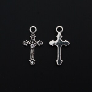 May include: Two silver-toned metal cross charms with a circular jump ring at the top. The left charm features a detailed embossed figure of a person on a cross, while the right charm displays a smooth, plain back side with a decorative cross shape.