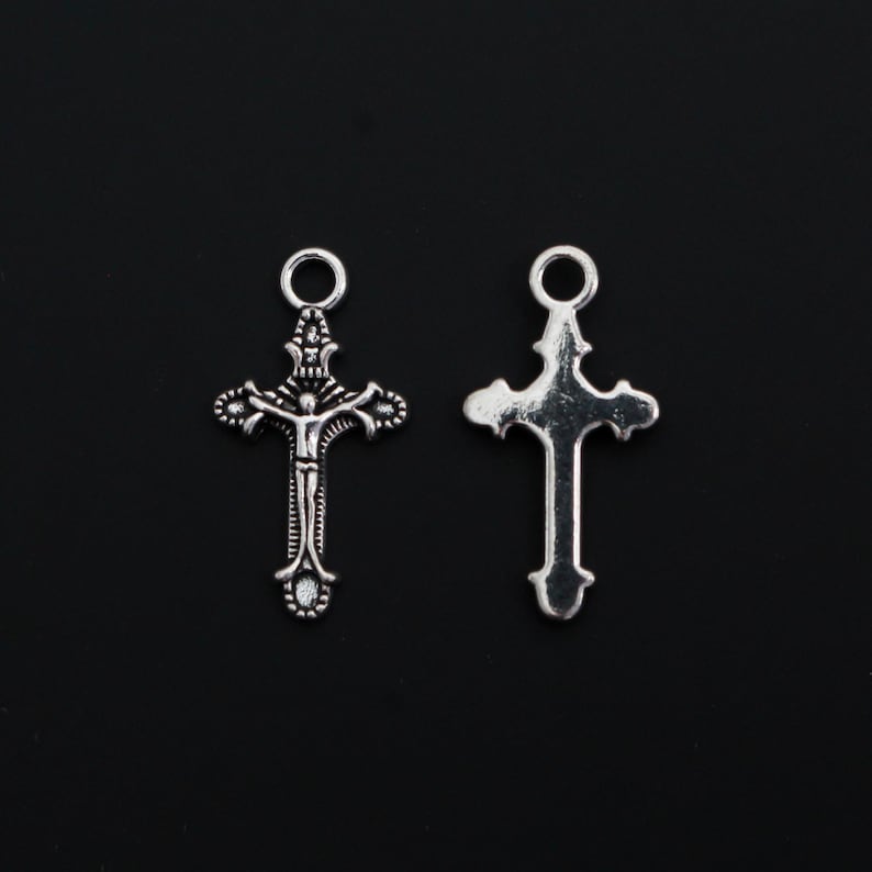 May include: Two silver-toned metal cross charms with a circular jump ring at the top. The left charm features a detailed embossed figure of a person on a cross, while the right charm displays a smooth, plain back side with a decorative cross shape.