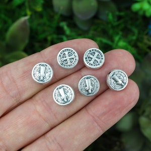 May include: A collection of small, round, silver-colored beads. Each bead features a detailed embossed design, including religious symbols and text. The beads are displayed against a blurred green background, suggesting a natural setting.
