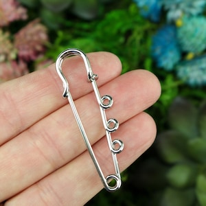 May include: A large, silver-colored safety pin with three decorative loops. The pin is held against a blurred background of green and blue foliage. The pin's design is simple and elegant, suitable for various crafting or fashion projects.
