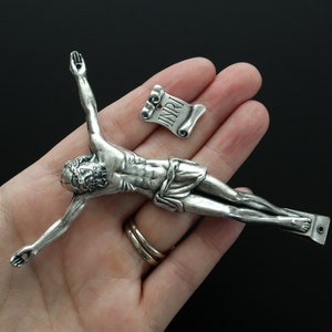 May include: A silver-toned metal crucifix featuring a detailed figure of Jesus on the cross with arms outstretched. A separate small silver-toned metal scroll piece is positioned above the head, clearly engraved with the letters INRI.