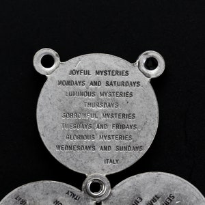 May include: A silver-coloured religious pendant with three circular sections. The top section has two holes and text that reads "JOYFUL MYSTERIES" and "MONDAYS AND SATURDAYS." The bottom sections have additional text. The word "ITALY" is also visible.