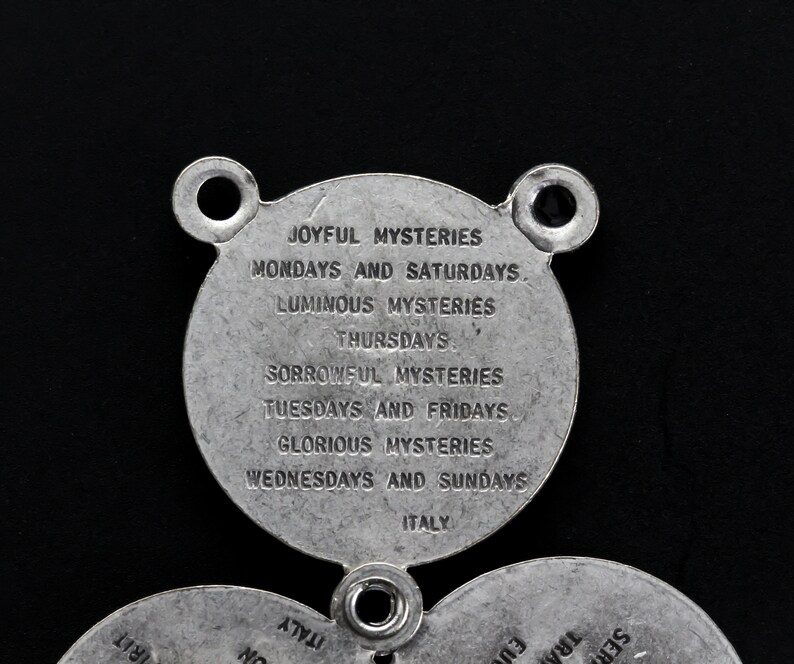 May include: A silver-coloured religious pendant with three circular sections. The top section has two holes and text that reads "JOYFUL MYSTERIES" and "MONDAYS AND SATURDAYS." The bottom sections have additional text. The word "ITALY" is also visible.