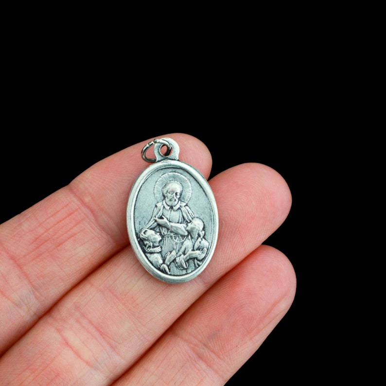 May include: Silver oval pendant featuring a religious image of Saint Anthony of Padua holding two children. The pendant has a loop at the top for attaching to a chain.