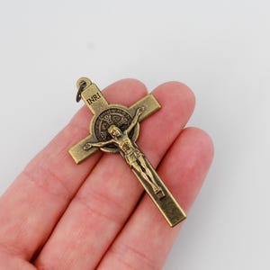 Op de afbeelding: Een bronzen kruisbeeld met de figuur van Jezus Christus. Het kruis heeft een kleine lus aan de bovenkant om op te hangen. De letters "INRI" zijn zichtbaar aan de bovenkant van het kruis. De figuur van Jezus bevindt zich in een kruisigingshouding.
