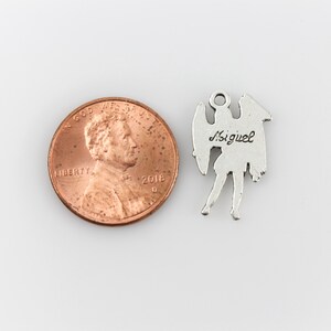May include: Silver charm of an angel with wings, standing on one leg. The back of the charm is engraved with the name "Miguel". The charm is shown next to a US penny for size comparison.