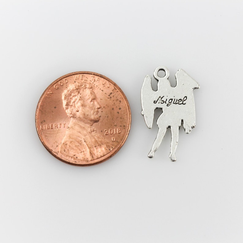 May include: Silver charm of an angel with wings, standing on one leg. The back of the charm is engraved with the name "Miguel". The charm is shown next to a US penny for size comparison.