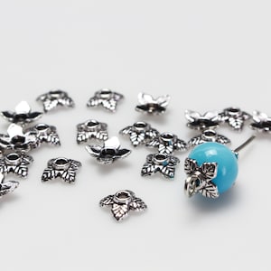 May include: Silver-toned metal bead caps with a leaf design. The caps are scattered on a white surface with a single turquoise bead in the center.