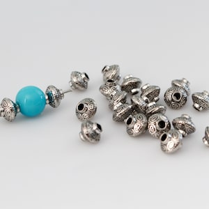 Puede incluir: Cuentas plateadas con un diseño ornamentado, dispersas sobre una superficie blanca. Una hebra presenta una cuenta turquesa brillante y detalles plateados. Adecuado para la creación de joyas.