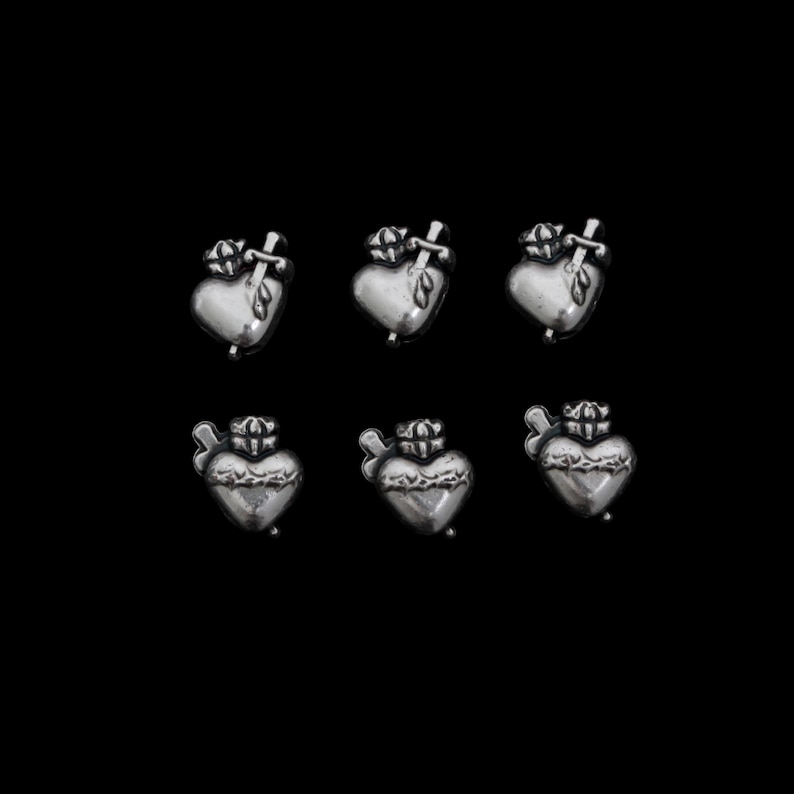 May include: Six silver-toned heart-shaped charms with a crown and a sword piercing the heart. The charms are arranged in two rows of three against a black background. The hearts have a detailed, textured design.