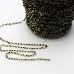 May include: A spool of antique brass-colored chain. The chain is made of small, oval links and is partially unraveled, with a length of chain extending from the spool. The chain is on a white surface.