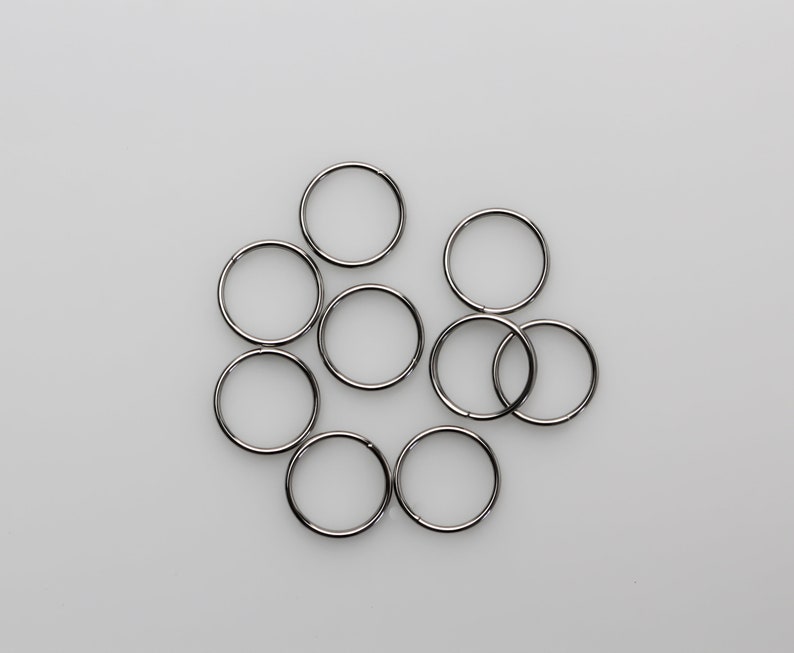 May include: A collection of nine silver-colored metal split rings, arranged in a loose cluster against a plain white background. The rings are circular and appear to be made of a shiny, reflective material.