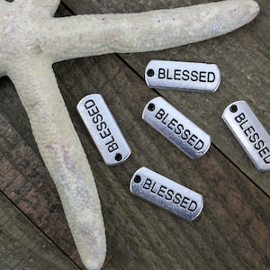 May include: Five silver metal tags with the word "BLESSED" engraved on them. The tags are arranged on a wooden surface with a white starfish.
