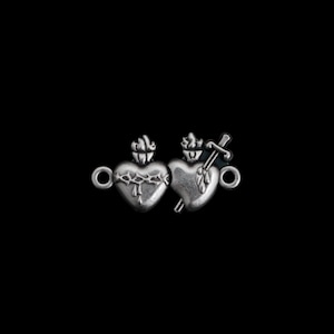 May include: Two silver heart charms with detailed designs. One heart has a crown of thorns, the other is pierced by a sword. Each charm has a loop for attaching to a chain or other jewellery component. The background is black.