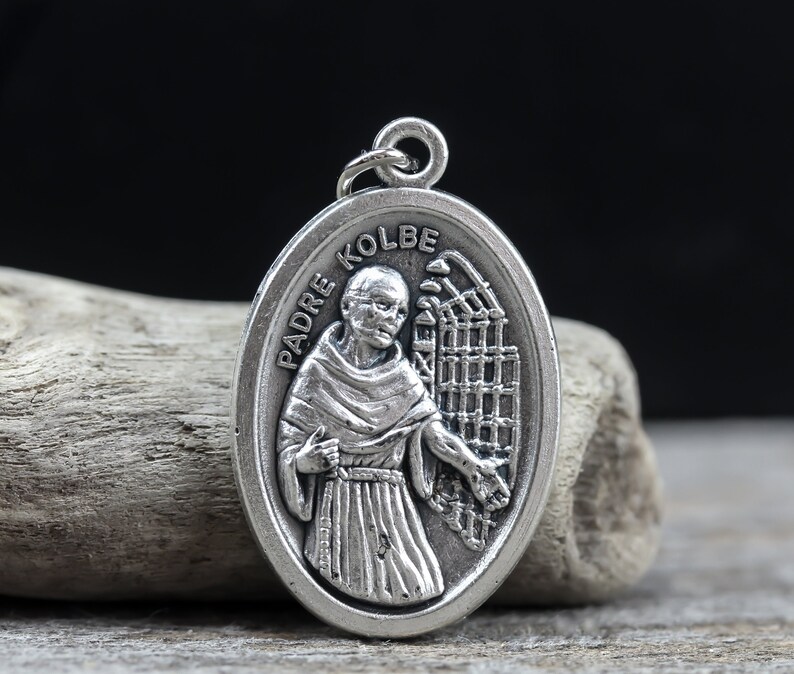 May include: Silver oval pendant with a depiction of Padre Kolbe, a Catholic saint, wearing a brown robe and holding a cross. The pendant features the words "Padre Kolbe" inscribed around the edge.