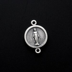May include: Silver charm featuring a depiction of the Virgin Mary with a halo, standing with her hands raised in prayer. The charm is round and has a detailed design.