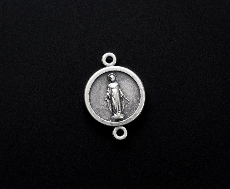 May include: Silver charm featuring a depiction of the Virgin Mary with a halo, standing with her hands raised in prayer. The charm is round and has a detailed design.