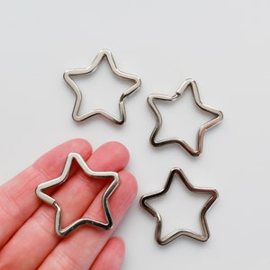 May include: Four silver star-shaped keychains. Three keychains are on a white surface, and one keychain is held in a hand.