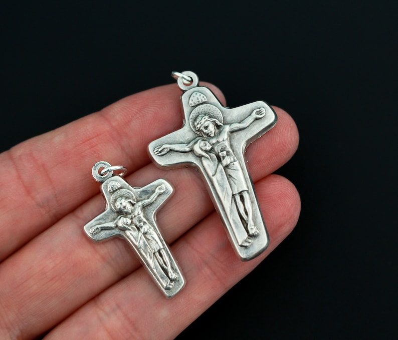 May include: Two silver crucifixes with a raised image of Jesus Christ on the cross. The larger crucifix is about 2 inches tall and the smaller crucifix is about 1 inch tall.