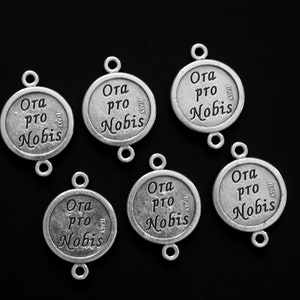May include: Six silver-toned metal charms with the words "Ora pro Nobis" and "Italy" engraved on them. The charms have two loops for attaching to a necklace or bracelet.