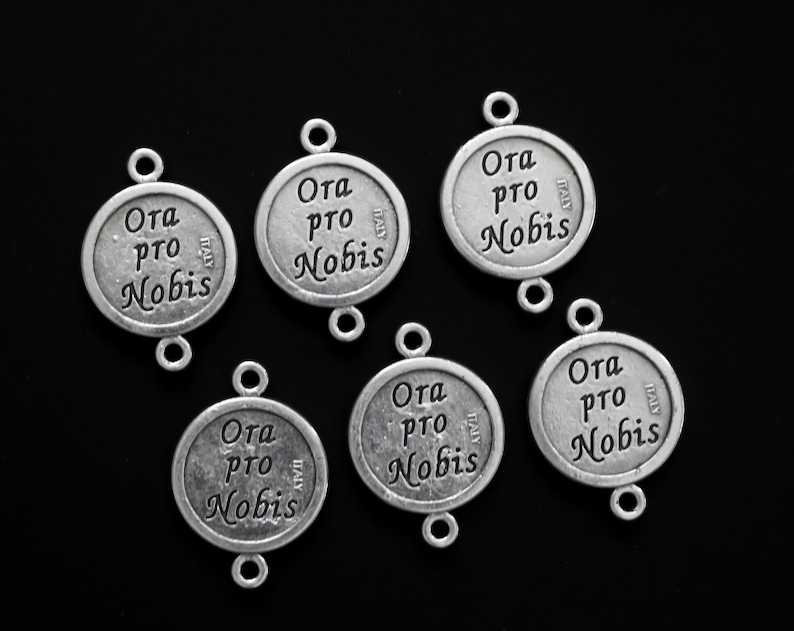 May include: Six silver-toned metal charms with the words "Ora pro Nobis" and "Italy" engraved on them. The charms have two loops for attaching to a necklace or bracelet.