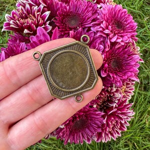 May include: A bronze-colored metal pendant with a geometric design, featuring a central circular space and four small loops. The pendant is held against a backdrop of vibrant, deep pink chrysanthemum flowers.