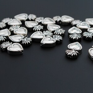 May include: A group of silver heart-shaped charms with a flame design. The charms are scattered on a black surface.