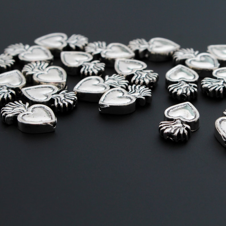 May include: A group of silver heart-shaped charms with a flame design. The charms are scattered on a black surface.