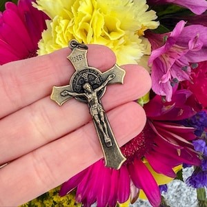 Op de afbeelding: Een kruisbeeld in bronskleur met de figuur van Jezus Christus. Het kruis heeft de inscriptie "INRI" bovenaan en wordt tegen een achtergrond van kleurrijke bloemen gehouden. Het kruisbeeld is een religieus item.